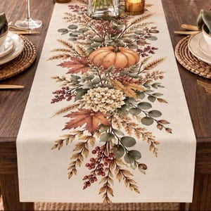 May include: A cream-colored table runner featuring a central botanical print of an orange pumpkin, beige hydrangea, brown maple leaves, green eucalyptus sprigs, and red berries arranged in a symmetrical vertical pattern along the fabric.