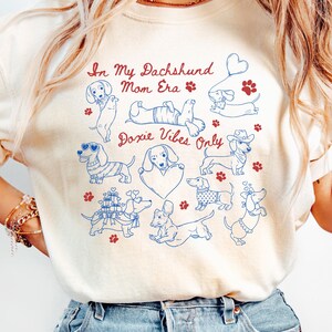 Ivory Comfort Colors® t-shirt featuring multiple dachshund illustrations with playful poses, hearts, and paw prints in a retro style design.