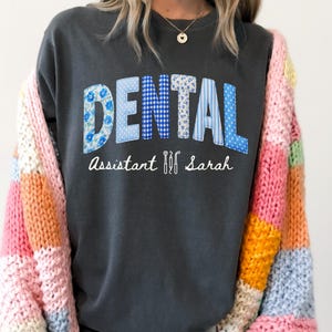 May include: Dark gray sweatshirt with the word "DENTAL" in patchwork letters, and the text "Assistant Sarah" below. The sweatshirt is paired with a colorful patchwork cardigan. The word "DENTAL" is made up of blue floral, gingham, and striped patterns.