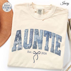 May include: An ivory-colored t-shirt featuring the word AUNTIE in a blue patchwork-style font. Below the text, the words Est. 2026 are printed in dark blue, separated by a small dark blue bow graphic. The shirt has a crew neckline and a label.