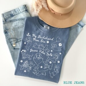 Blue Jeans Comfort Colors® t-shirt featuring multiple dachshund illustrations with playful poses, hearts, and paw prints in a retro style design.