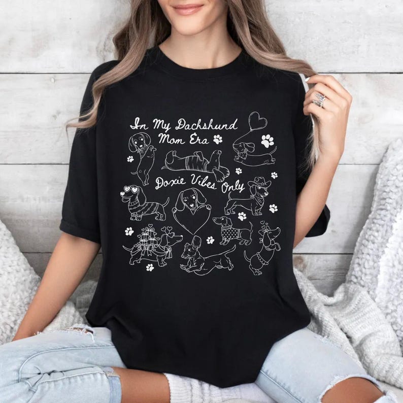 Black Comfort Colors® t-shirt featuring multiple dachshund illustrations with playful poses, hearts, and paw prints in a retro style design.