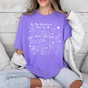 Violet Comfort Colors® t-shirt featuring multiple dachshund illustrations with playful poses, hearts, and paw prints in a retro style design.