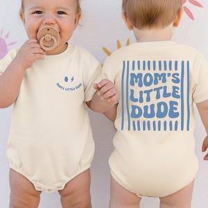May include: Two babies in cream-coloured onesies. The front of the onesie has the text "Mom's Little Dude" with a lightning bolt graphic. The back of the onesie has the text "MOM'S LITTLE DUDE" in blue. One baby is facing forward with a dummy.