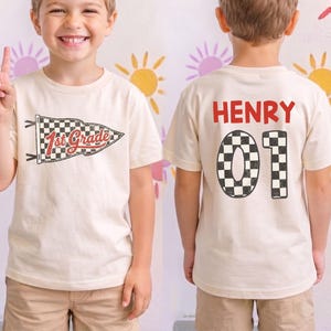 May include: A cream-colored short-sleeve t-shirt featuring a black and white checkered pennant graphic with the text 1st Grade on the front. The back displays the name Henry in red letters above the large checkered numbers 01 in black and white.