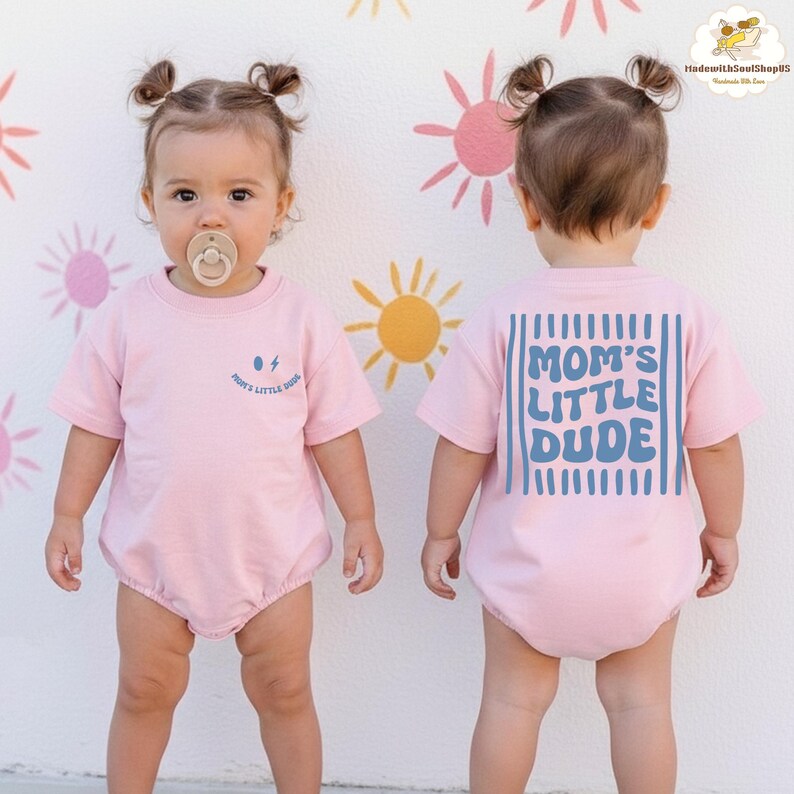 May include: A pink baby romper with short sleeves. The front has the text "Mom's Little Dude" with a lightning bolt graphic. The back of the romper features the text "MOM'S LITTLE DUDE" in blue, with vertical blue lines.