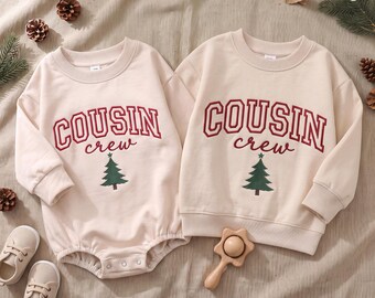Puede incluir: Un pelele de bebé de manga larga en color beige y una sudadera beige a juego, ambos con el texto rojo COUSIN y el texto rojo en cursiva crew sobre un gráfico de un árbol de Navidad verde. Las prendas tienen cuellos redondos y puños acanalados.