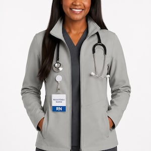 May include: A light gray zip-up jacket with a high collar, worn over a dark gray V-neck scrub top and matching pants. A stethoscope is draped around the neck, and a white ID badge with the text REGISTERED NURSE RN is clipped to the jacket.
