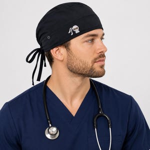 May include: A solid black surgical scrub cap featuring a small embroidered white ninja icon on the side. The cap has a wide band, a rounded crown, and two long black fabric ties at the back for an adjustable fit. The fabric is smooth and matte.