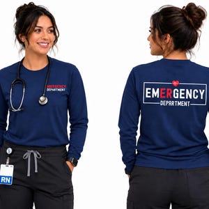 May include: A navy blue long-sleeve shirt featuring the text "EMERGENCY DEPARTMENT" on the front left chest. The back of the shirt displays a large white rectangular frame containing the text "EMERGENCY DEPARTMENT" with a red heart icon.