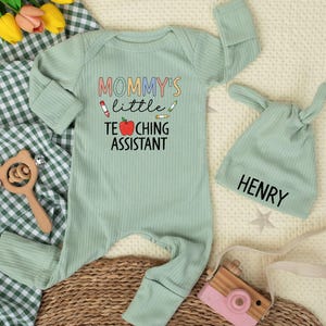 May include: A light green baby outfit set with the text "MOMMY'S little TEACHING ASSISTANT" and a matching hat with the name "HENRY". The set includes a long-sleeved onesie and a hat with bunny ears.
