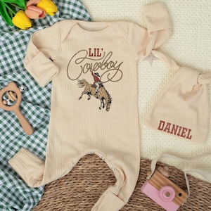 May include: A cream-colored baby outfit set featuring a long-sleeved onesie with "Lil' Cowboy" text and a cowboy graphic, and a matching knotted hat with the name "DANIEL". The set is displayed with a wooden rattle and a toy camera.