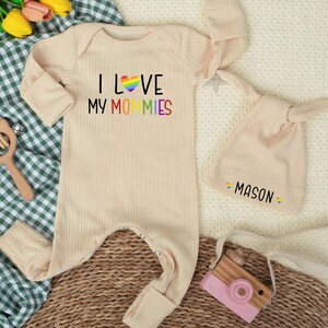 May include: A beige ribbed baby onesie with the text "I LOVE MY MOMMIES" in black and rainbow colors. A matching knotted hat with the name "MASON" and rainbow accents is included. A wooden toy camera and rattle are also in the image.