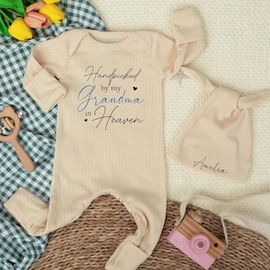 May include: A beige baby onesie and matching knotted hat set. The onesie has the text "Handpicked by my Grandma in Heaven" in black and blue script. The hat is personalized with the name "Amelia". A wooden toy camera and a rattle are also in the image.
