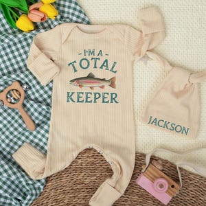 May include: A beige ribbed baby onesie with the text "I'M A - TOTAL KEEPER" and a fish graphic. A matching knotted hat with the name "JACKSON" is next to the onesie. A wooden toy camera and a wooden rattle are also in the image.