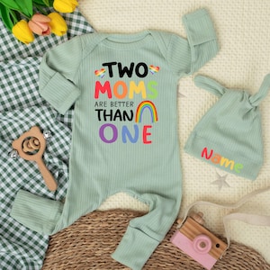 May include: Sage green baby onesie and matching hat. The onesie features the text "TWO MOMS ARE BETTER THAN ONE" with a rainbow graphic. The hat has the word "Name" in rainbow colors. A wooden toy and a toy camera are also in the image.