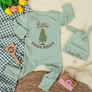 May include: A mint green baby outfit set featuring a long-sleeved onesie and a matching knotted hat. The onesie has the text "Little Hiking Buddy" with a tree graphic. The hat is embroidered with the name "Sadie Mae".