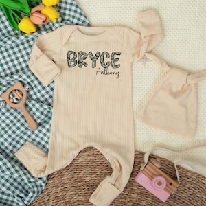 May include: A beige ribbed baby onesie with the name "Bryce" in camouflage print and the name "Anthony" in black script. A matching knotted hat and a wooden toy are next to the onesie. The onesie has long sleeves and foot coverings.