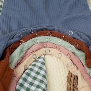 May include: A close-up of several baby onesies in various colours, including blue, brown, green, pink, and beige. Each onesie has a ribbed texture and metal snap closures. A green and white checked fabric and a woven basket are visible.
