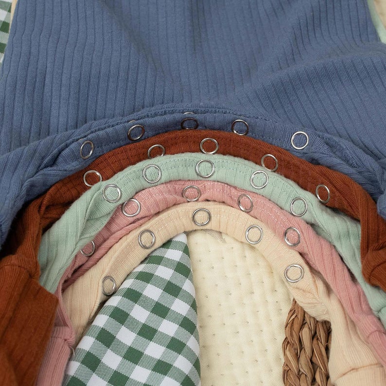 May include: A close-up of several baby onesies in various colours, including blue, brown, green, pink, and beige. Each onesie has a ribbed texture and metal snap closures. A green and white checked fabric and a woven basket are visible.