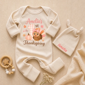May include: A cream-colored ribbed long-sleeve baby romper and matching knotted hat. The romper features a graphic of a turkey with a pink bow, a pumpkin, and the text Amelia's 1st Thanksgiving. The hat is embroidered with the name Amelia.
