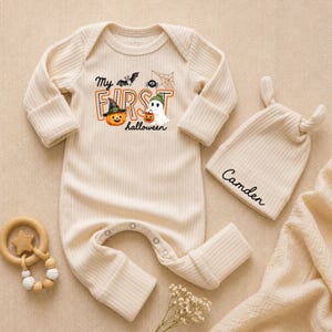 May include: A cream-colored ribbed long-sleeve baby romper with a graphic reading My First Halloween featuring a pumpkin, ghost, bat, and spider. A matching cream ribbed knotted hat with the name Camden printed in black script is included.