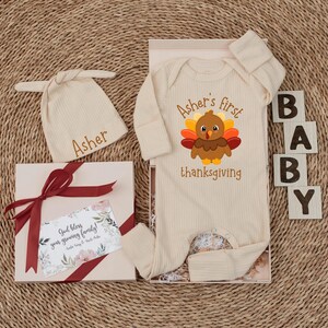 May include: A cream-colored ribbed baby bodysuit and matching knotted hat set. The bodysuit features a graphic of a turkey with the text Asher's first thanksgiving. A gift box includes a card with the text God bless your growing family.