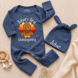 May include: A navy blue ribbed long-sleeve romper with a graphic of a turkey and the text "Asher's first thanksgiving" in white, paired with a matching knotted beanie featuring the name "Asher" in white text, both made of soft fabric.