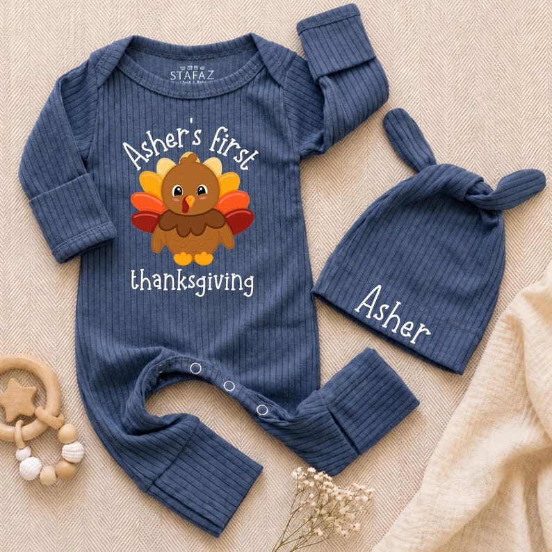 May include: A navy blue ribbed long-sleeve romper with a graphic of a turkey and the text "Asher's first thanksgiving" in white, paired with a matching knotted beanie featuring the name "Asher" in white text, both made of soft fabric.