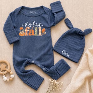 May include: A dark blue ribbed long-sleeve baby romper with a graphic print that reads My first fall featuring a pumpkin and autumn leaves. The set includes a matching dark blue ribbed baby hat with the name Olivia printed in white script text.