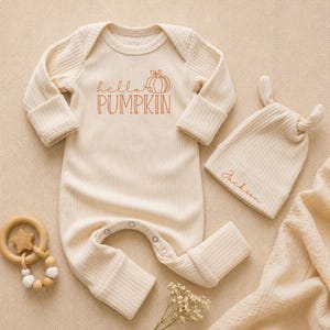 May include: A cream-colored ribbed long-sleeve romper with a snap-button crotch and the text "hello PUMPKIN" in orange, paired with a matching ribbed beanie featuring the embroidered name "Jackson" and a wooden star-shaped teething ring.