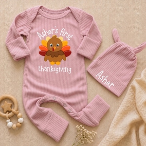 May include: A dusty pink ribbed long-sleeve baby romper with a graphic of a turkey and the text "Asher's first thanksgiving" and a matching dusty pink baby beanie with two small ears and the name "Asher" embroidered in white text.