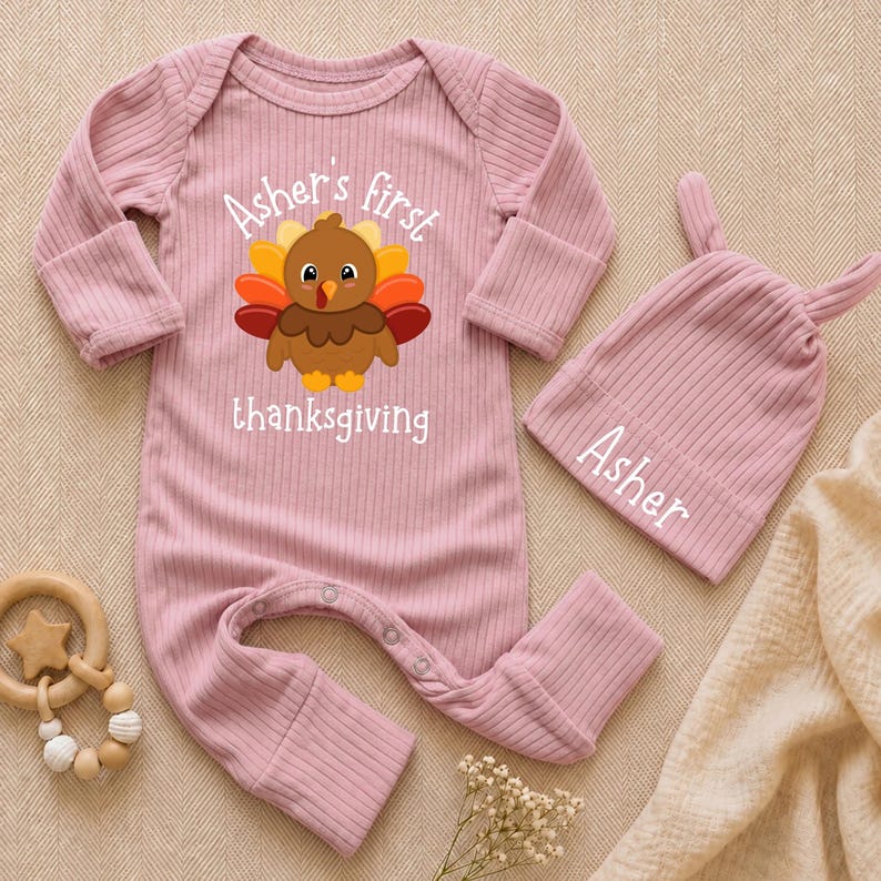 May include: A dusty pink ribbed long-sleeve baby romper with a graphic of a turkey and the text "Asher's first thanksgiving" and a matching dusty pink baby beanie with two small ears and the name "Asher" embroidered in white text.