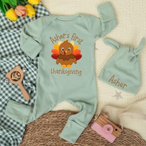 May include: A sage green ribbed long-sleeve baby romper and matching knotted hat. The romper features a graphic of a brown turkey with colorful feathers and the text "Asher's first thanksgiving". The hat is embroidered with the name "Asher".