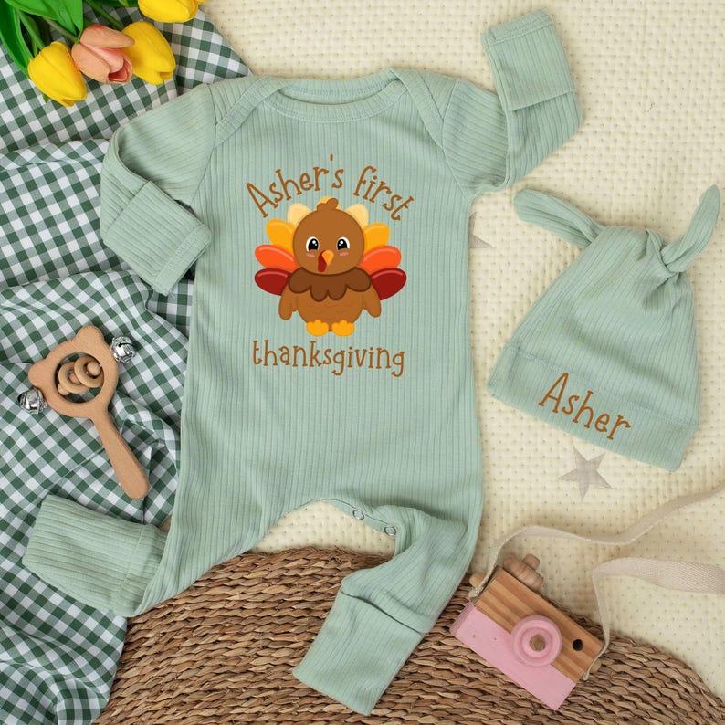 May include: A sage green ribbed long-sleeve baby romper and matching knotted hat. The romper features a graphic of a brown turkey with colorful feathers and the text "Asher's first thanksgiving". The hat is embroidered with the name "Asher".
