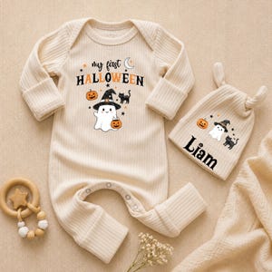 May include: A cream-colored ribbed long-sleeve romper and matching knotted hat set. The romper features the text "my first HALLOWEEN" with a ghost wearing a witch hat, a pumpkin, and a black cat. The hat is personalized with the name "Liam".