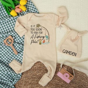 May include: A cream-coloured baby onesie with a fishing-themed graphic and the text "IS IT TOO SOON TO ASK FOR A FISHING POLE?". Includes a matching knotted hat with the name "CAMDEN" and a wooden toy camera. The outfit is ribbed.
