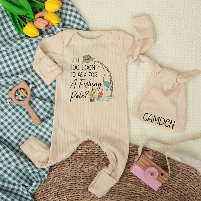 May include: A cream-coloured baby onesie with a fishing-themed graphic and the text "IS IT TOO SOON TO ASK FOR A FISHING POLE?". Includes a matching knotted hat with the name "CAMDEN" and a wooden toy camera. The outfit is ribbed.