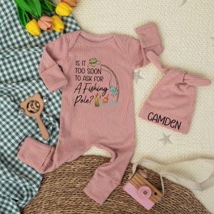 May include: A pink ribbed baby onesie with the text "Is it too soon to ask for a fishing pole?" and a matching hat with the name "Camden". A wooden toy camera and rattle are also in the image.