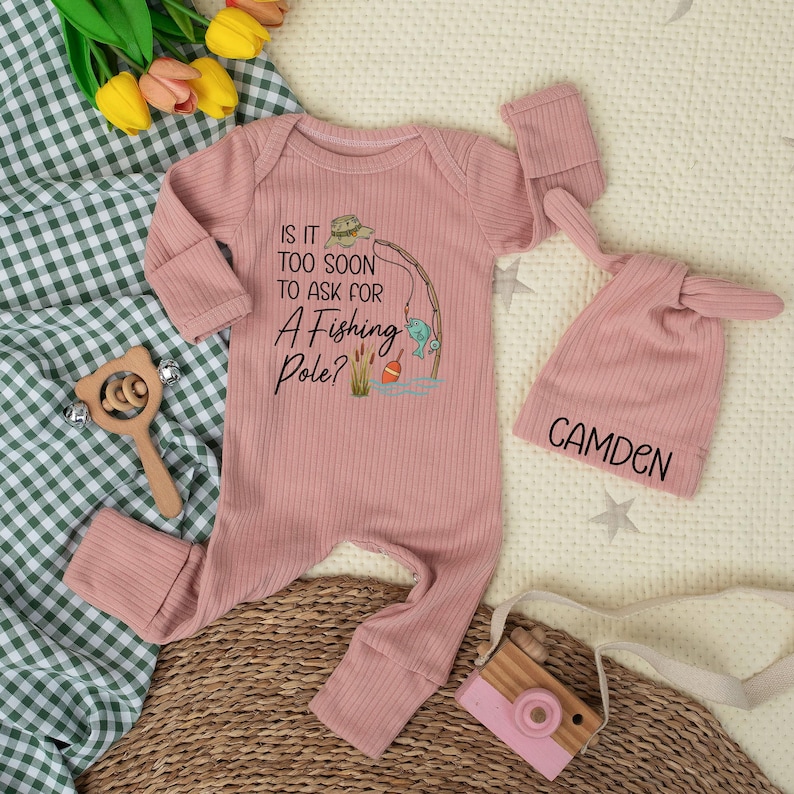 May include: A pink ribbed baby onesie with the text "Is it too soon to ask for a fishing pole?" and a matching hat with the name "Camden". A wooden toy camera and rattle are also in the image.