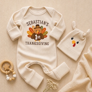 May include: A cream-colored ribbed long-sleeve baby bodysuit with a graphic of a turkey wearing a pilgrim hat and the text "SEBASTIAN'S 1st THANKSGIVING". Included is a matching cream ribbed beanie with a turkey face and a wooden teething ring.