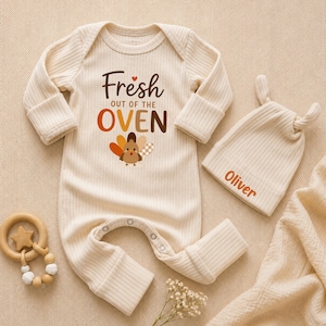 May include: A cream-colored ribbed long-sleeve baby romper with the text Fresh out of the oven and a turkey graphic. A matching cream-colored knotted baby hat features the name Oliver in orange text. A wooden star-shaped baby rattle is included.