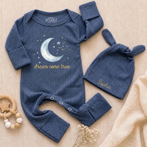 May include: A dark blue ribbed long-sleeve romper with a crescent moon and star graphic, the text "dream come true," and a matching knotted hat with the embroidered name "Sophia." The romper features a snap-button closure at the crotch area.