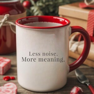 Puede incluir: Taza de cerámica color crema con borde y asa rojos. La taza tiene el texto "Less noise. More meaning." en la parte delantera. La taza está sobre una superficie de madera con detalles rojos y una cuchara.