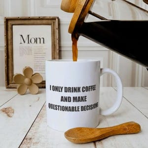 Puede incluir: Taza de cerámica blanca con asa negra y la frase "But First, Coffee" en negro. La taza se muestra sobre un fondo borroso, sugiriendo un ambiente acogedor.
