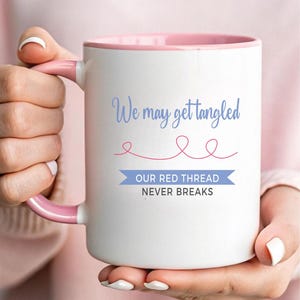 Puede incluir: Taza de cerámica blanca con interior y asa rosas. La taza presenta el texto "We may get tangled" en azul claro, con un diseño de hilo rojo y las palabras "OUR RED THREAD NEVER BREAKS" en una pancarta azul.