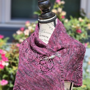 May include: A knitted shawl in shades of deep pink and black, draped over a mannequin. The shawl features a delicate openwork design and is secured with a silver-toned pin. The mannequin has the words "DE PORTE" printed on it.