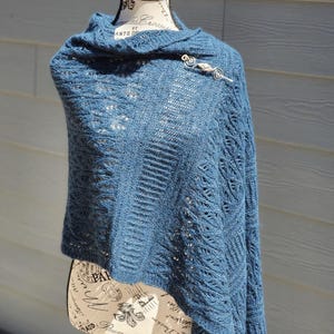 Può includere: Uno scialle blu lavorato a maglia drappeggiato su un manichino. Lo scialle è fissato con una spilla argentata. Il motivo a maglia presenta dettagli traforati e strutturati. Il manichino ha un top nero e un busto color crema.