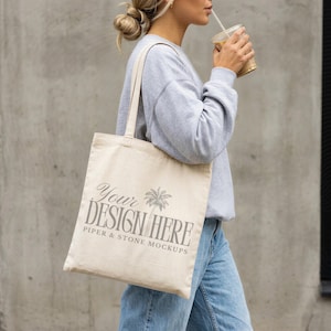 Puede incluir: Un bolso de tela beige con el texto YOUR DESIGN HERE y PIPER & STONE MOCKUPS junto a un gráfico de palmera. El bolso tiene dos asas y se lleva al hombro, combinado con una sudadera gris claro y unos pantalones vaqueros de color azul.