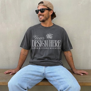 Könnte beinhalten: Ein anthrazitfarbenes Kurzarm-T-Shirt mit dem weißen Schriftzug "YOUR DESIGN HERE" und "PIPER & STONE MOCKUPS" sowie einer Palmen-Grafik. Das T-Shirt wird mit einer hellblauen Jeans, einer beigen Baseballkappe und einer schwarzen Sonnenbrille kombiniert.